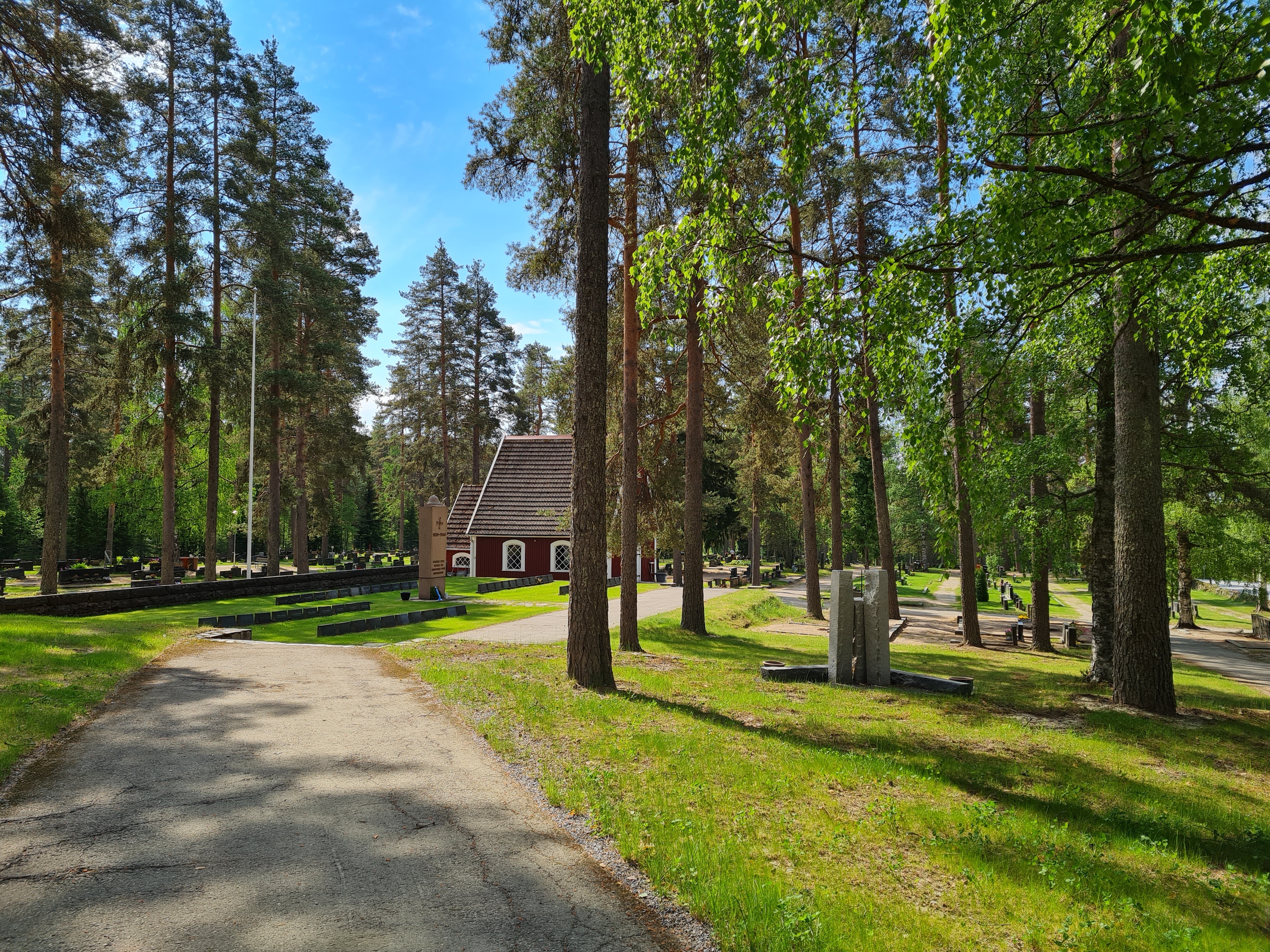 Kesäinen hautausmaa, jossa mäntyjä ja lipputanko. Sankarihautausmaa ja punainen hirsinen vanha kappeli